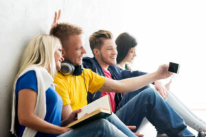 Group of happy students being on a break taking selfie. One of students is doing prank. Focus on a happy girl and boy in headphones. Background is blurry.